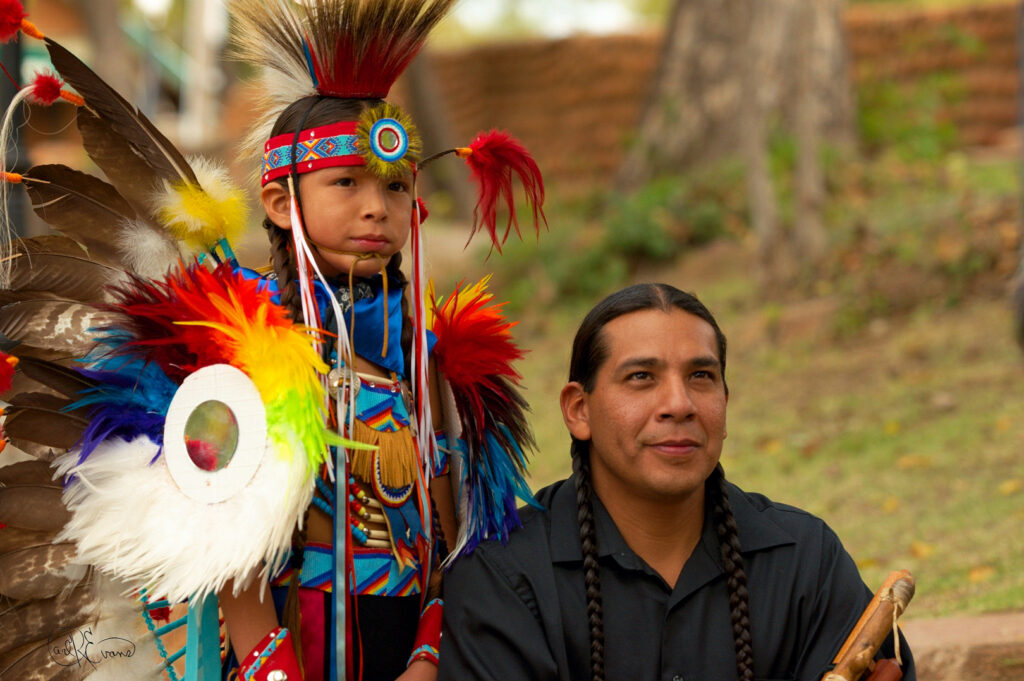 A young Comanche dancer stands with his father, a musician holding a flute.
