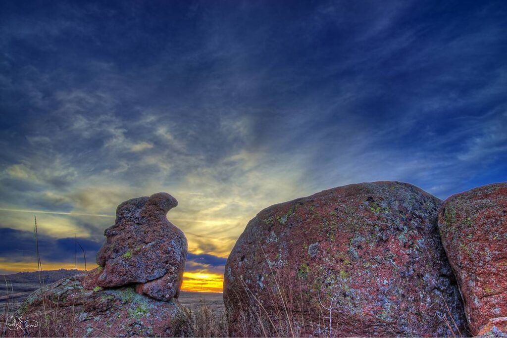 Red granite boulders frame a sunset view in the Wichita Mountains, Oklahoma, with wispy clouds overhead.
