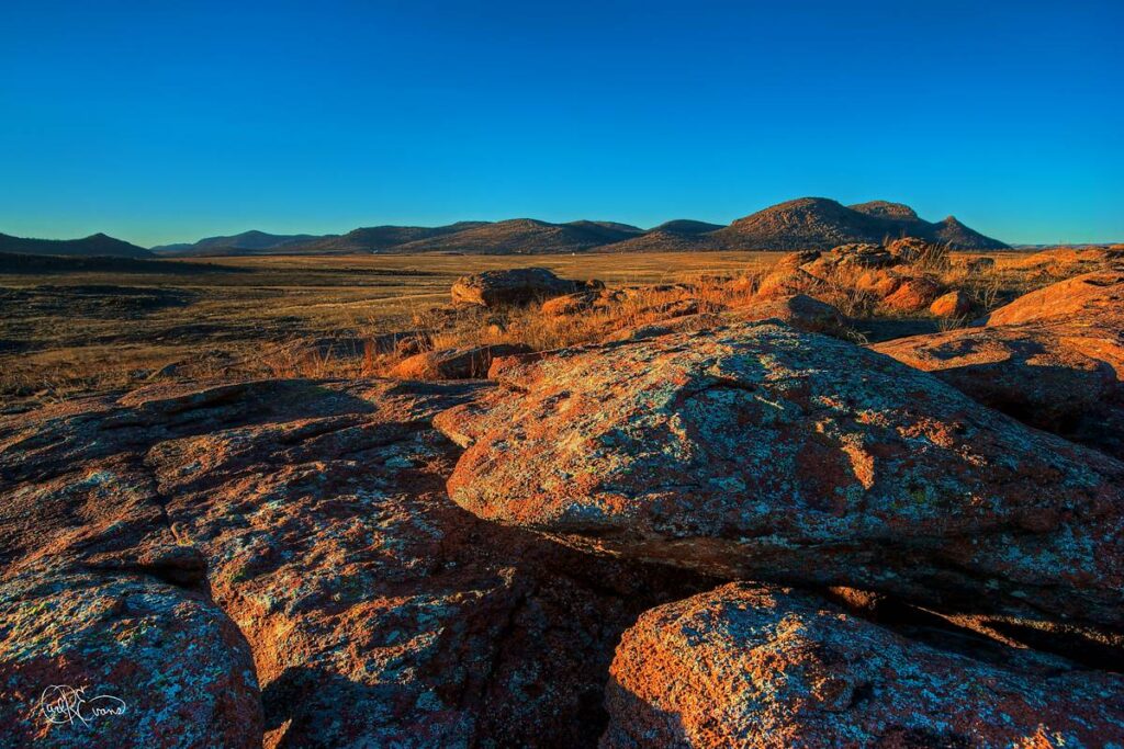Red granite boulders and a prairie landscape stretch towards distant hills under a clear blue sky.
