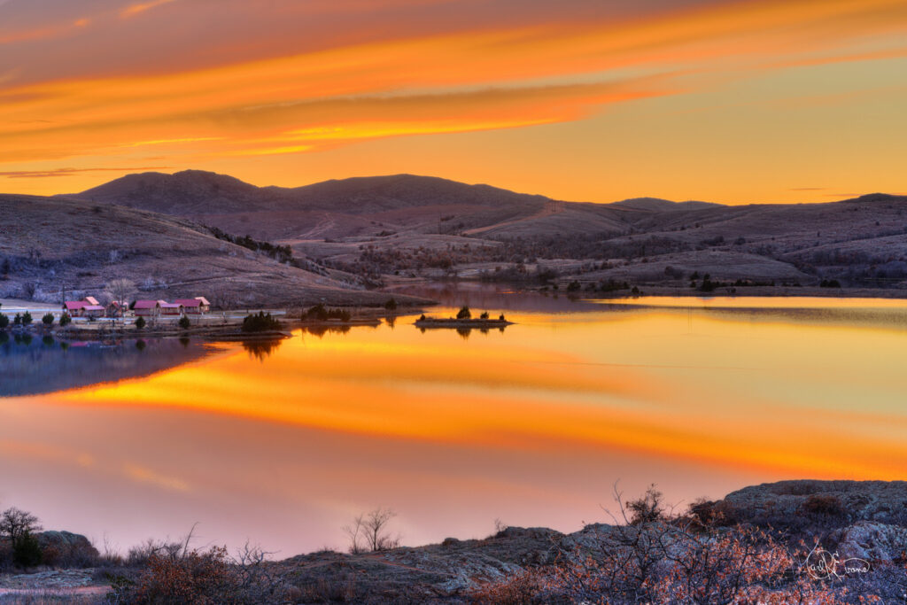 Sunset over Lake Elmer Thomas in Oklahoma, showing golden light reflecting off the still water and framed by the Wichita Mountains. A peaceful rural landscape.
