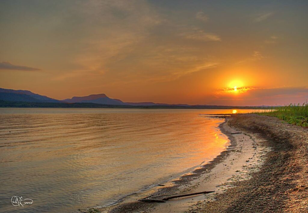 A serene sunset over Lake Lawtonka captures the tranquil beauty of a summer evening. The warm light reflects on the water's surface, creating a peaceful scene. This image showcases the natural splendor of Oklahoma's lakes