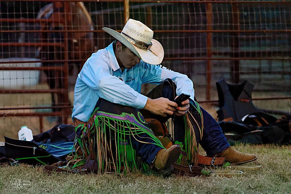 A young cowboy sits on grassy ground behind rodeo chutes, looking down at a cellphone during a hot, late summer evening.